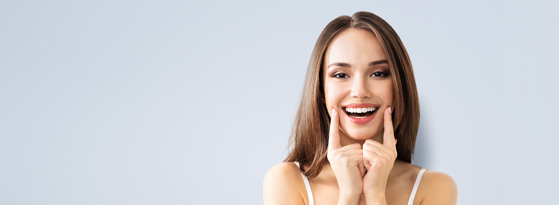 The image is a photograph of a woman with light skin, smiling at the camera. She appears to be in her late twenties or early thirties and has long hair. Her eyes are looking directly at the camera, and she is holding up her index finger near her mouth as if she s making a point or emphasizing something. The background is plain and light-colored, which suggests that this could be a stock photo used for various purposes such as advertising, personal branding, or lifestyle content.