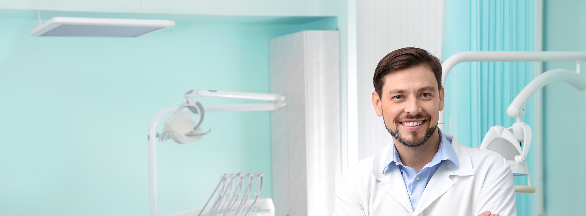 The image shows a smiling man in a dental office, standing behind a counter with medical equipment and wearing a white coat.