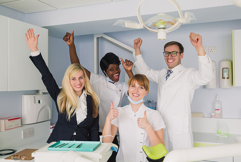 A group of five individuals, possibly dental professionals, in a dental office setting, celebrating with raised hands and thumbs up.
