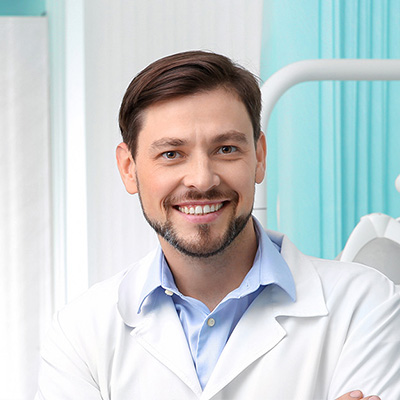 A smiling man in a white lab coat stands confidently in front of a dental chair, suggesting his role as a dentist.