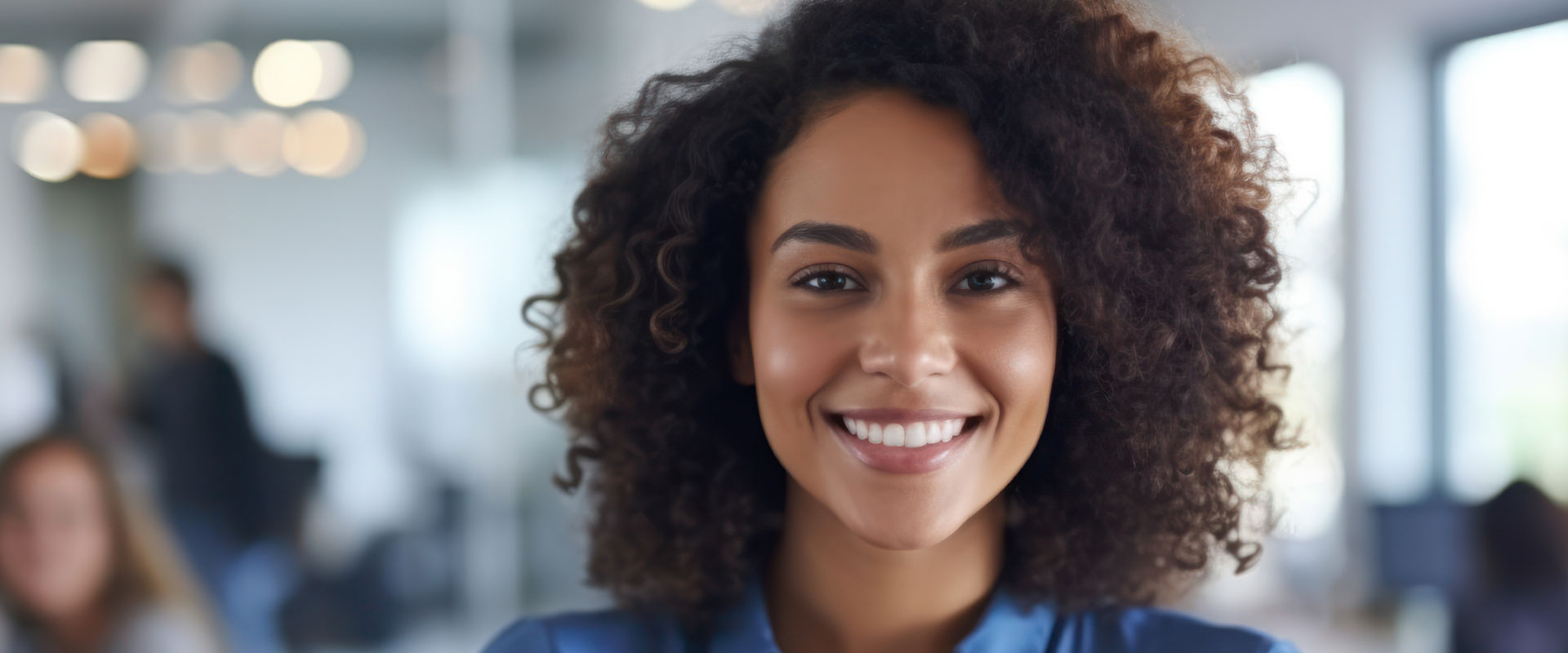 This is a photograph of a woman with a smile, wearing a blue shirt and standing in an office environment.