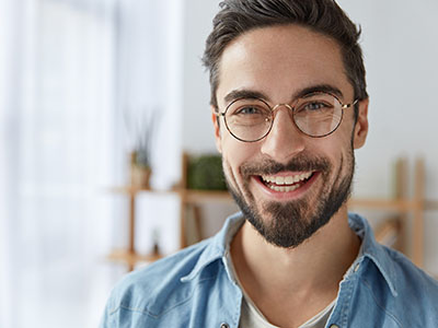 A smiling man with a beard and glasses, wearing a blue denim shirt, stands in front of a white wall.